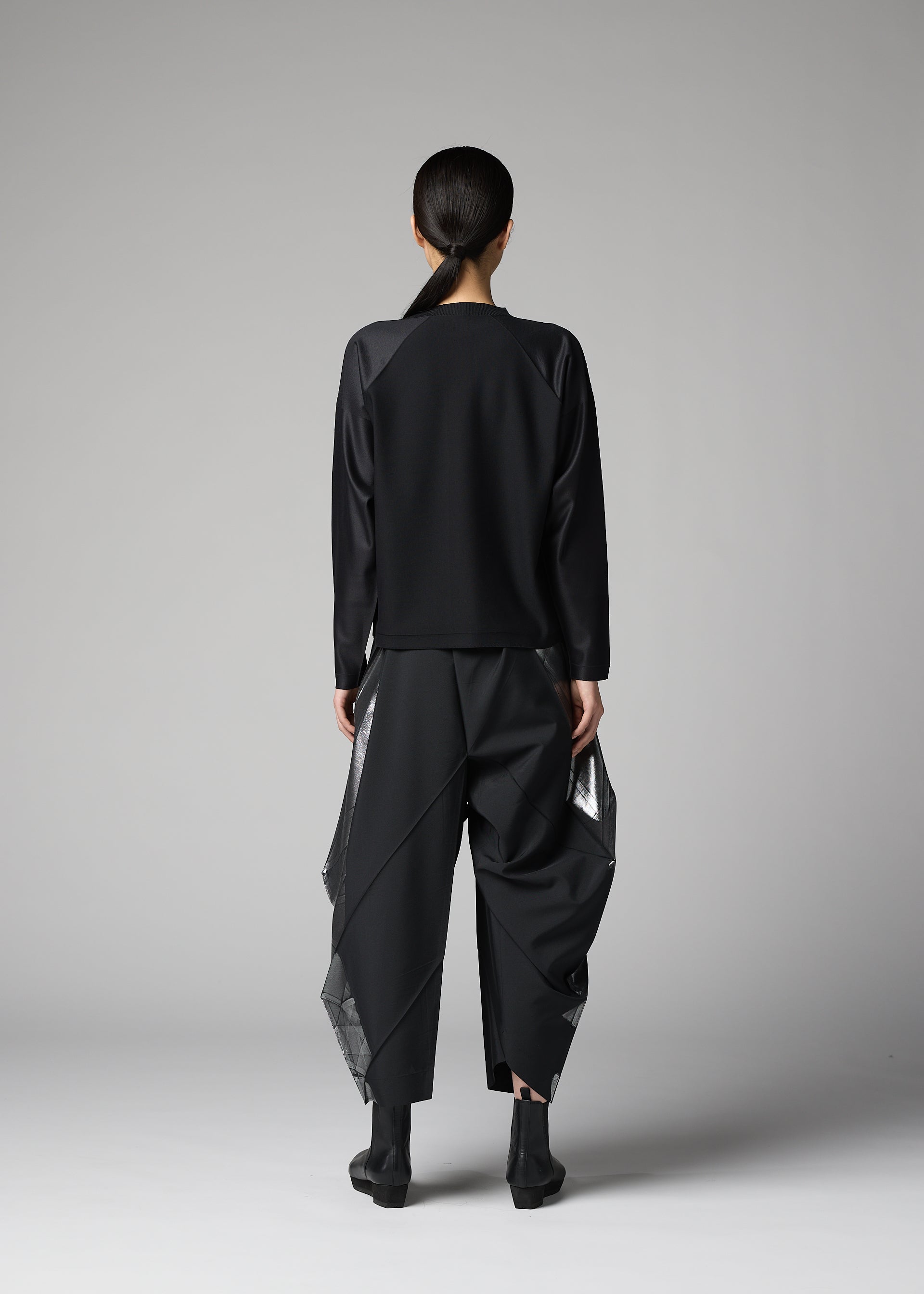 Zoomed view A person with long dark hair tied back stands facing away in a plain studio, wearing the F-KN black top and wide structured pants with shiny angular panels by 132 5. ISSEY MIYAKE, paired with black ankle boots.