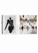 An open ISSEY MIYAKE (Limited Collectors Edition) book by ISSEY MIYAKE BOOKS displays a black-and-white photograph of a model in an intricate outfit on the left page, alongside a color image of models suspended midair in an ornate gallery on the right page, reflecting Issey Miyake’s experimental design philosophy.