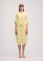 A woman with straight, light brown hair and bangs wears ISSEY MIYAKE’s LINKAGE set—a textured, pale yellow knit skirt and matching top with wavy scalloped edges, made from recycled polyester—against a plain background. She pairs it with sandals.