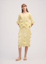 A woman poses against a plain background wearing the LINKAGE skirt and top set by ISSEY MIYAKE, featuring pale yellow textured knits with ruffled edges and matching flat sandals, her straight light brown hair with bangs completing the look.