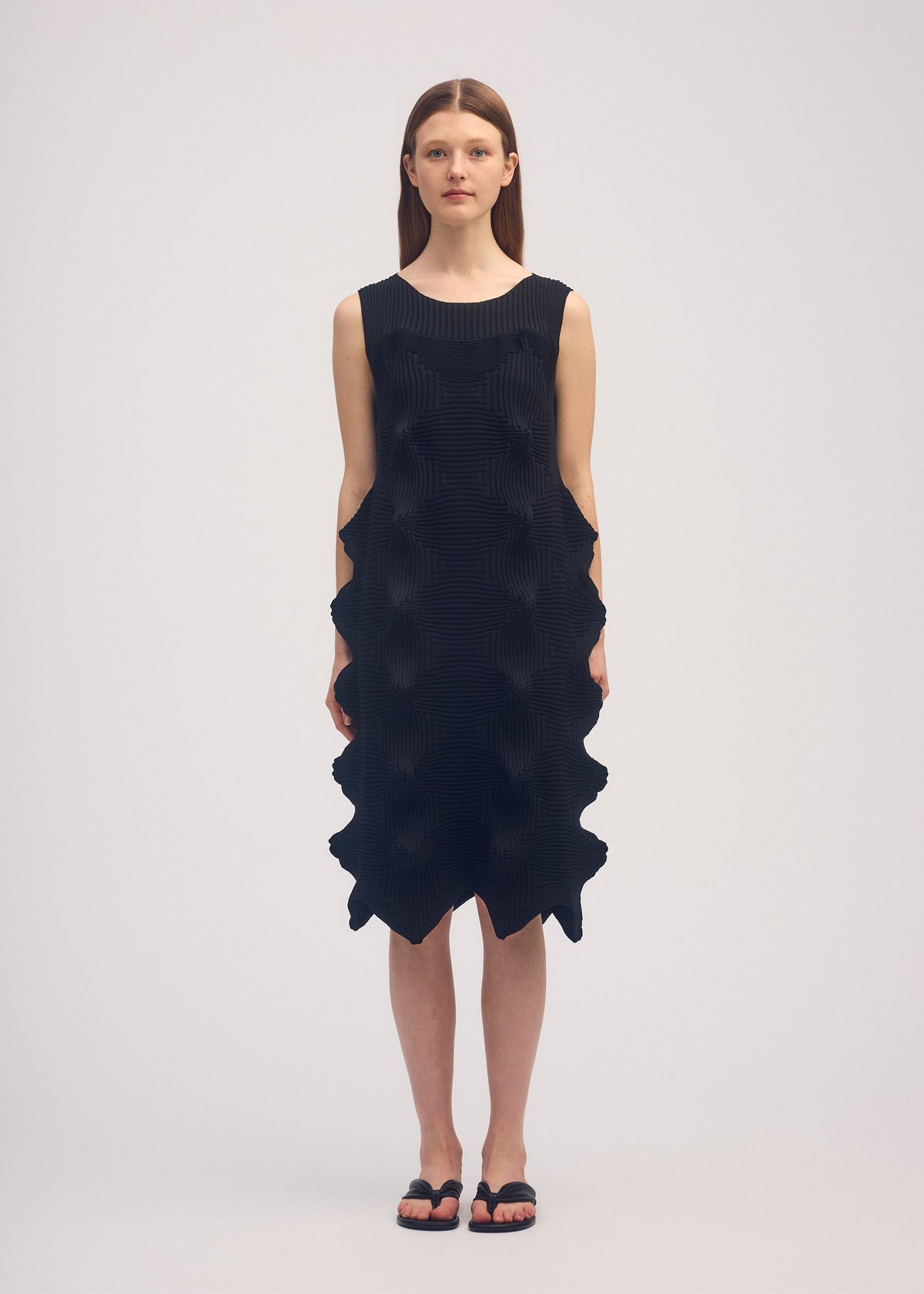 A woman stands facing forward in the LINKAGE dress by ISSEY MIYAKE—a sleeveless, knee-length black knit dress with a geometric textured pattern, jagged edges, and a bold three-dimensional silhouette. She has straight brown hair and wears black sandals. view 2 of 3
