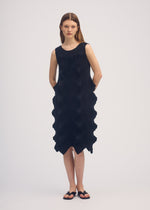 A woman poses against a plain background wearing the LINKAGE sleeveless knee-length black knit dress by ISSEY MIYAKE, featuring a wavy textured side pattern and a three-dimensional silhouette, styled with simple black sandals.
