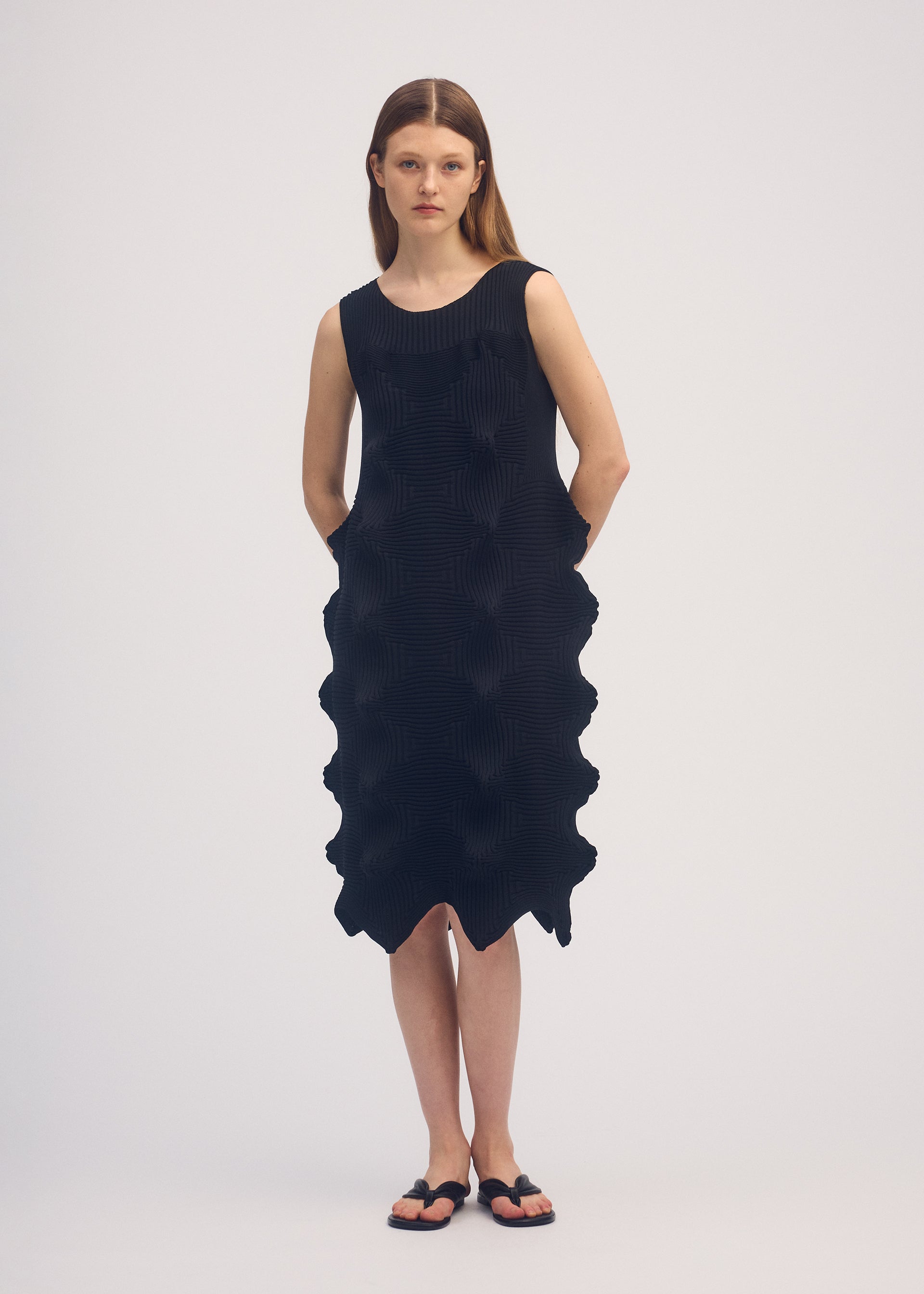 Zoomed view A woman poses against a plain background wearing the LINKAGE sleeveless knee-length black knit dress by ISSEY MIYAKE, featuring a wavy textured side pattern and a three-dimensional silhouette, styled with simple black sandals.