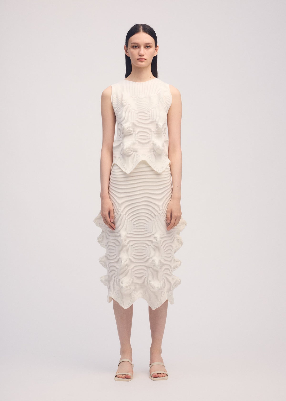 A woman faces the camera wearing ISSEY MIYAKE’s LINKAGE knit sleeveless top and matching knee-length skirt with sculpted wavy details, straight dark hair, neutral expression, and white sandals. view 3 of 4