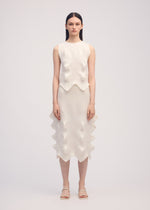 A woman faces the camera wearing ISSEY MIYAKE’s LINKAGE knit sleeveless top and matching knee-length skirt with sculpted wavy details, straight dark hair, neutral expression, and white sandals.