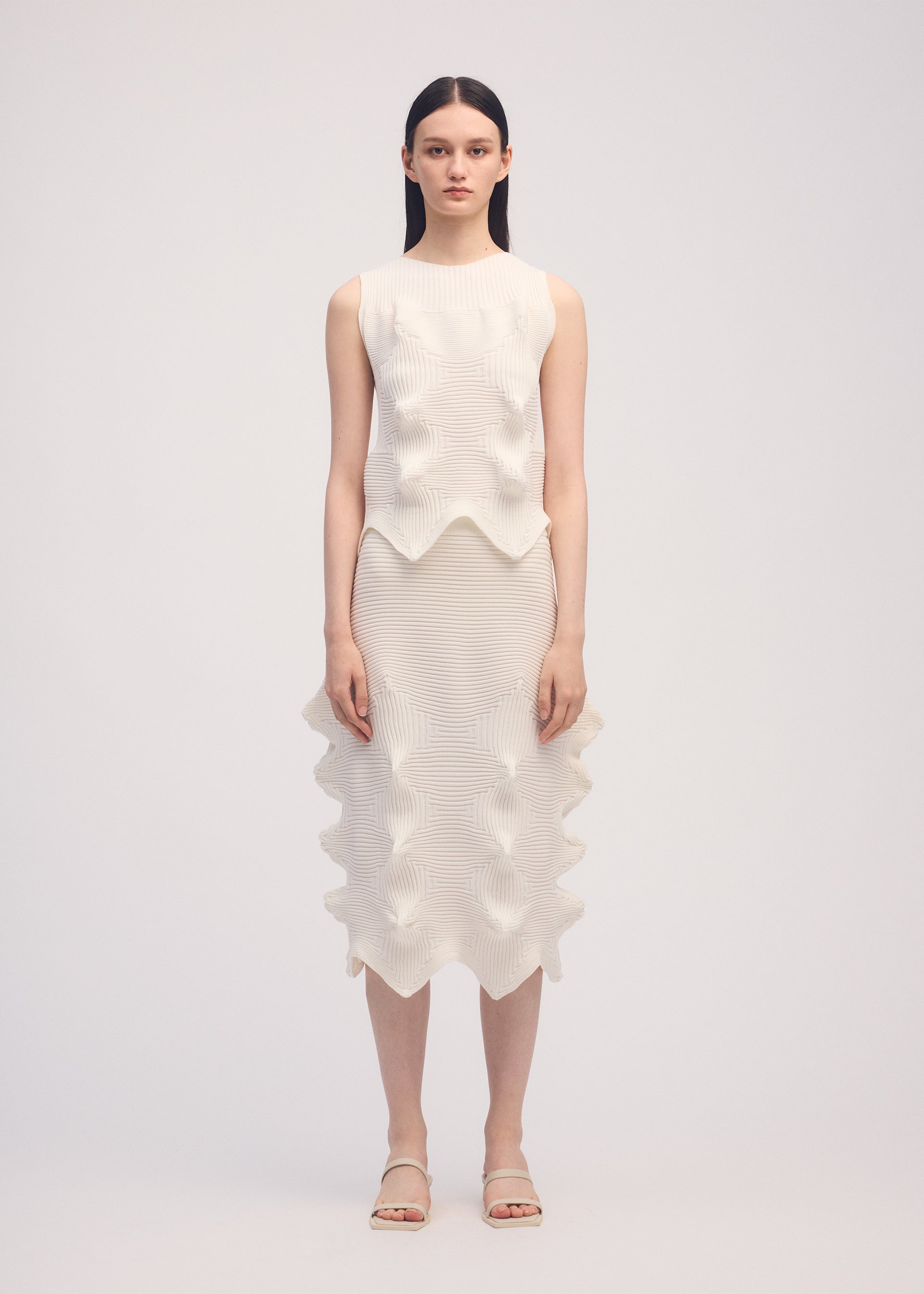 Zoomed view A woman faces the camera wearing ISSEY MIYAKE’s LINKAGE knit sleeveless top and matching knee-length skirt with sculpted wavy details, straight dark hair, neutral expression, and white sandals.