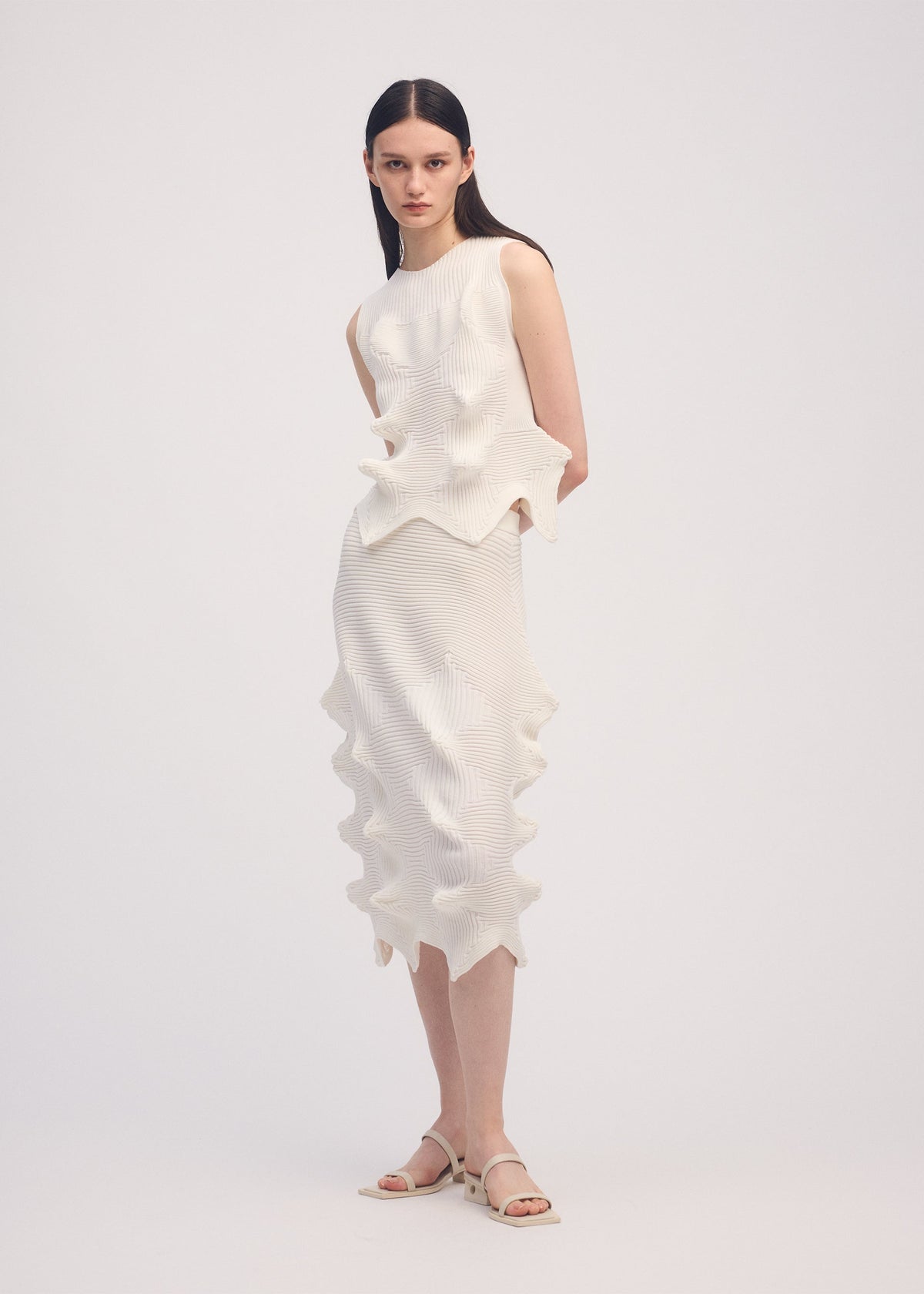 A woman with straight dark hair stands against a plain background wearing ISSEY MIYAKE's LINKAGE: a sleeveless, textured white dress featuring wavy, sculptural details and a three-dimensional silhouette. She pairs it with white sandals. view 4 of 4
