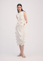 A woman with straight dark hair stands against a plain background wearing ISSEY MIYAKE's LINKAGE: a sleeveless, textured white dress featuring wavy, sculptural details and a three-dimensional silhouette. She pairs it with white sandals.