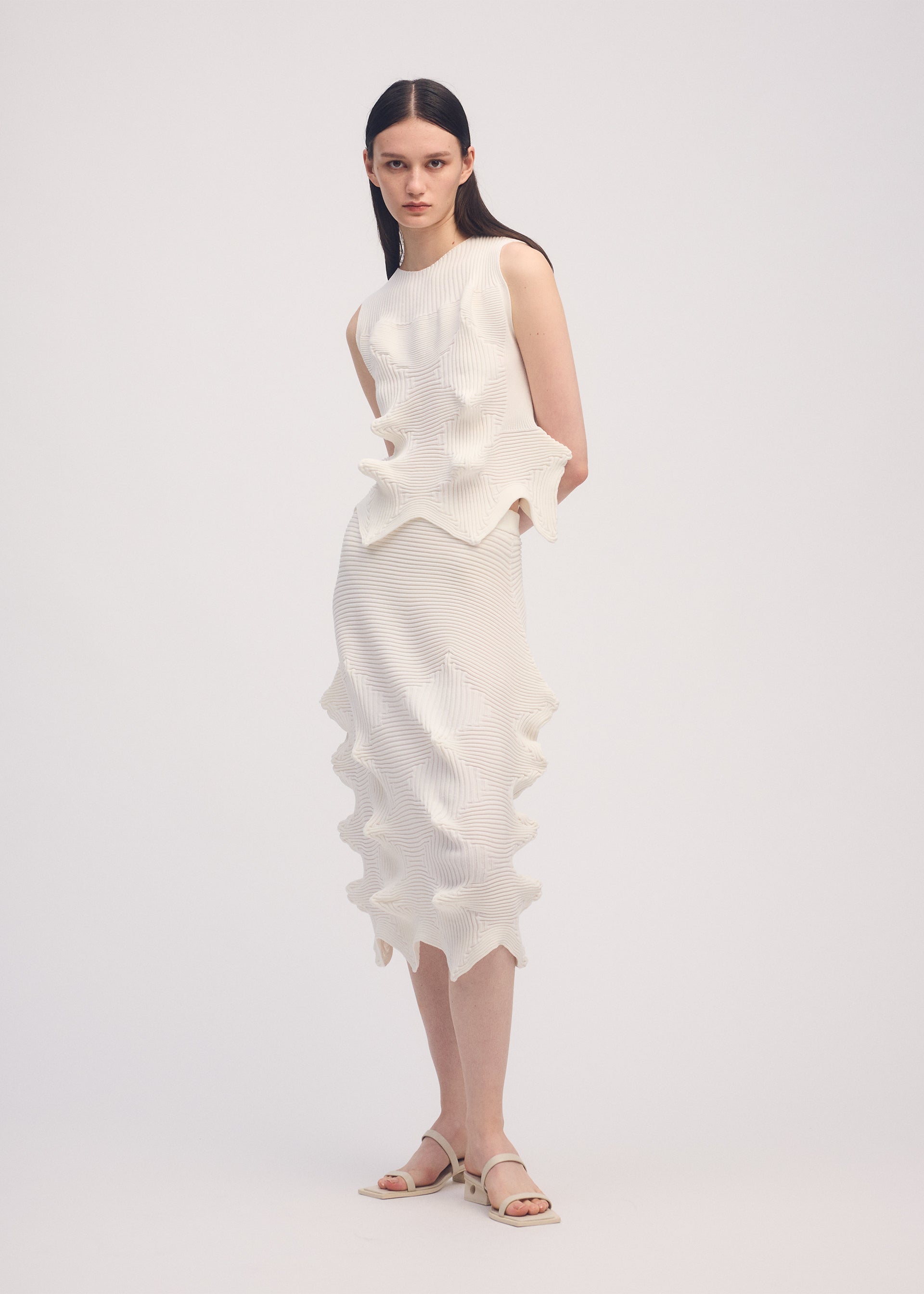 Zoomed view A woman with straight dark hair stands against a plain background wearing ISSEY MIYAKE's LINKAGE: a sleeveless, textured white dress featuring wavy, sculptural details and a three-dimensional silhouette. She pairs it with white sandals.