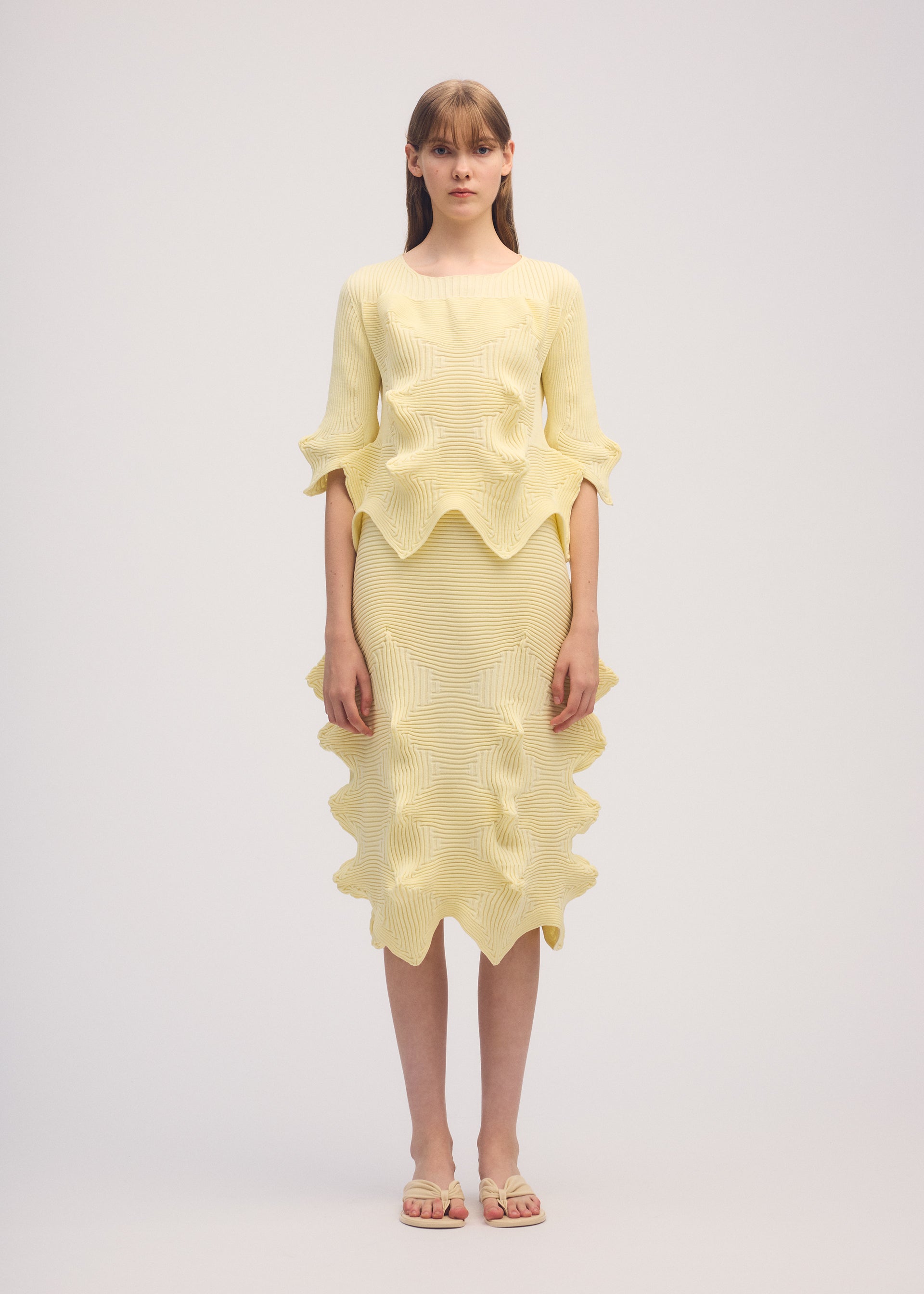 Zoomed view A woman faces forward in ISSEY MIYAKE's LINKAGE set: a pale yellow knit short-sleeved top and matching skirt with sculpted wavy edges. She pairs it with beige sandals, her straight light brown hair with bangs, against a plain light background.