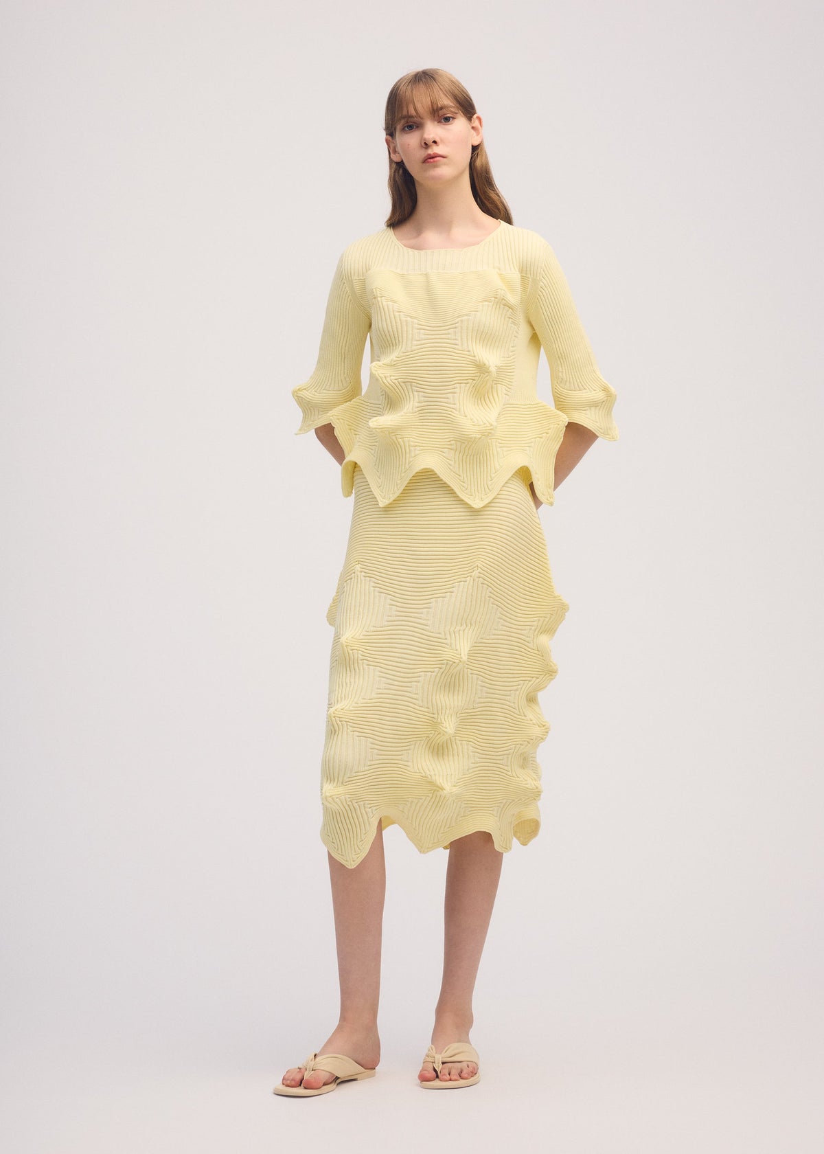 A woman with straight light brown hair and bangs wears ISSEY MIYAKE's LINKAGE: a light yellow, textured two-piece outfit with a 3D silhouette and matching sandals, standing against a plain background. view 3 of 3