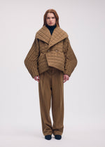 A person wearing a tan PLEATED GRID padded coat with handmade pleats on the wide sleeves over matching trousers and a black mock-neck top.
