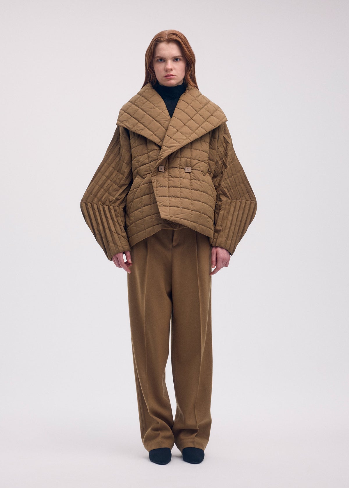 Zoomed view A person wearing a tan PLEATED GRID padded coat with handmade pleats on the wide sleeves over matching trousers and a black mock-neck top.