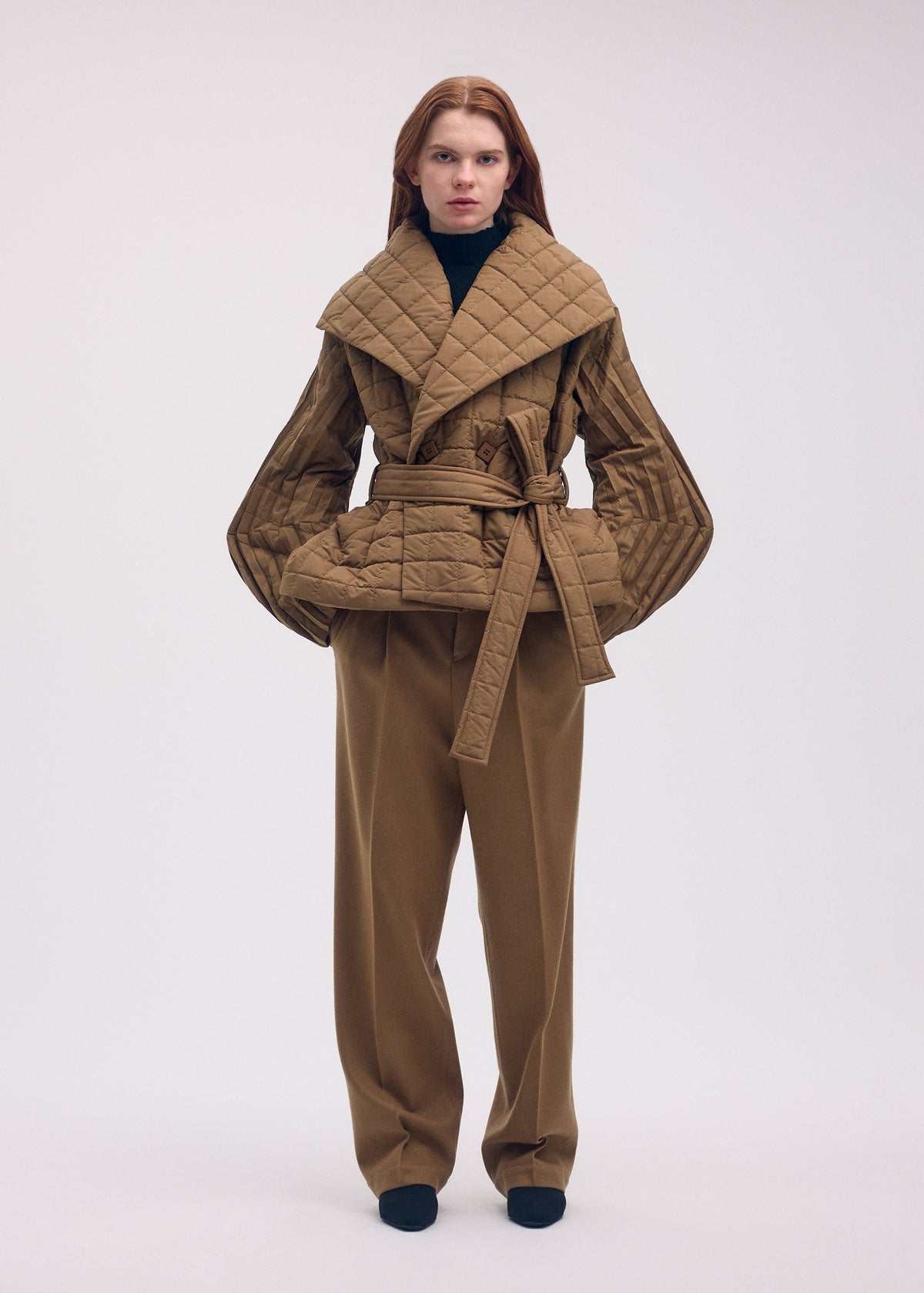 A person wears an ISSEY MIYAKE PLEATED GRID tan, padded coat with a square pattern, wide collar, and tie waist, paired with matching wide-leg trousers and a black turtleneck. view 3 of 4