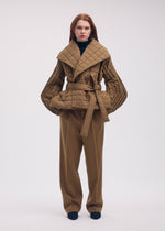 A person wears an ISSEY MIYAKE PLEATED GRID tan, padded coat with a square pattern, wide collar, and tie waist, paired with matching wide-leg trousers and a black turtleneck.