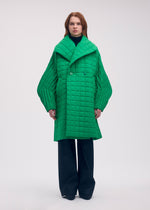 A person wears a bright green, oversized, long-padded PLEATED GRID coat by ISSEY MIYAKE, featuring wide pleated sleeves, a large lapel, and a distinctive square pattern.