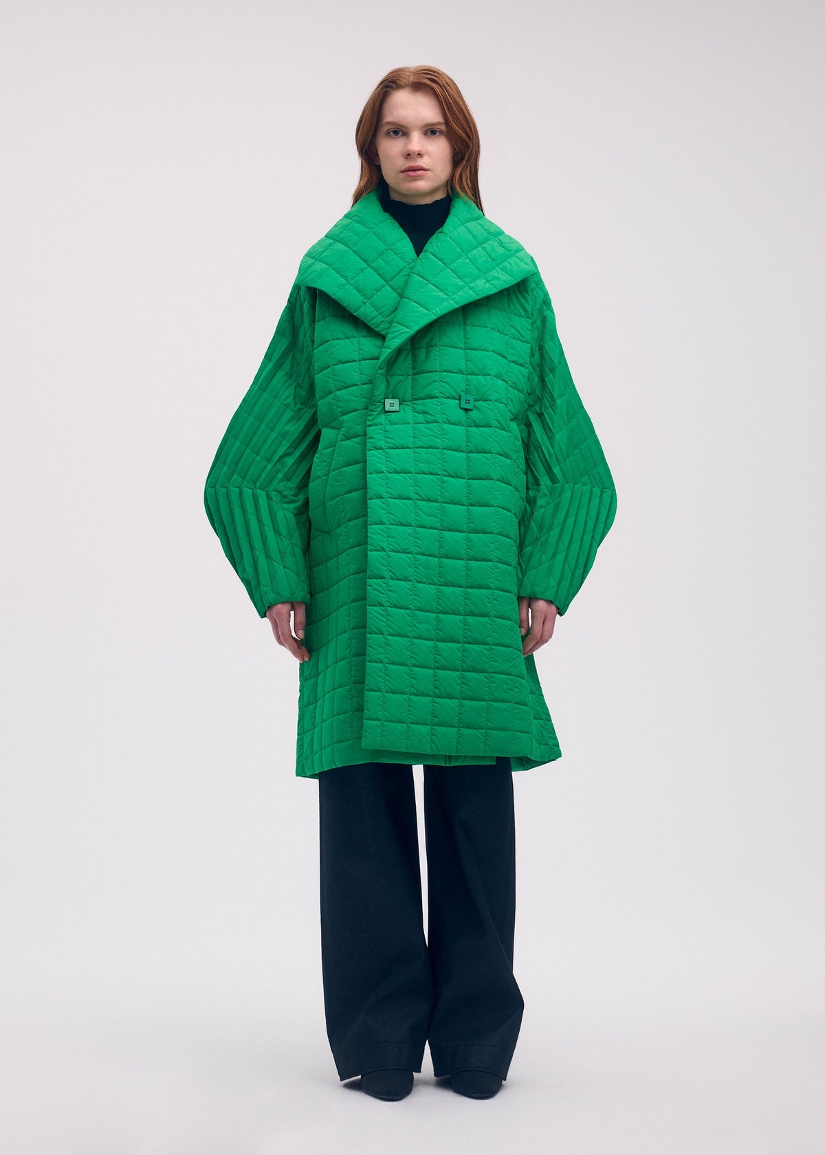 Zoomed view A person wears a bright green, oversized, long-padded PLEATED GRID coat by ISSEY MIYAKE, featuring wide pleated sleeves, a large lapel, and a distinctive square pattern.