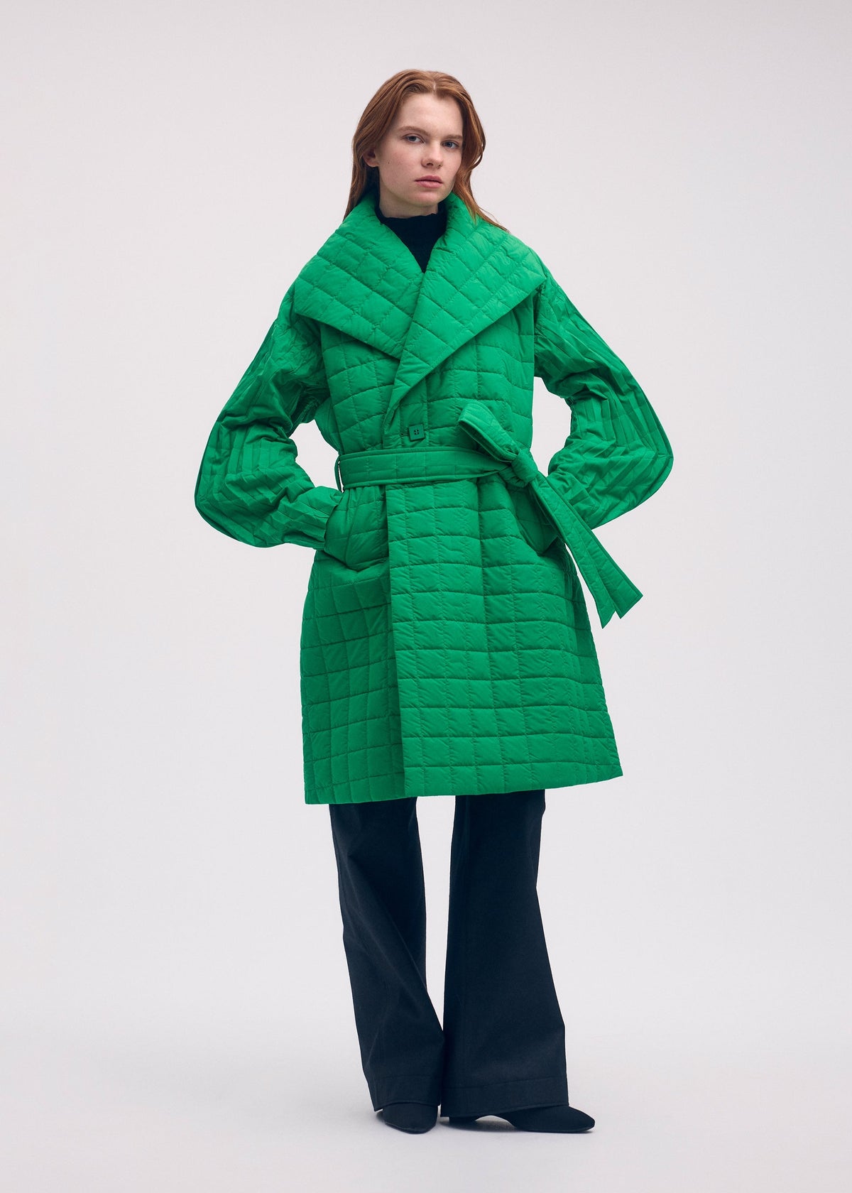 Zoomed view A person wears an ISSEY MIYAKE PLEATED GRID bright green, square-patterned, quilted, belted coat with a large collar and pleated sleeves over dark trousers.