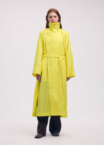 A person faces forward in the ISSEY MIYAKE AERIAL STEEL: a long, bright yellow lightweight taffeta coat with high collar and belt, worn over dark wide-leg pants, set against a plain light background.