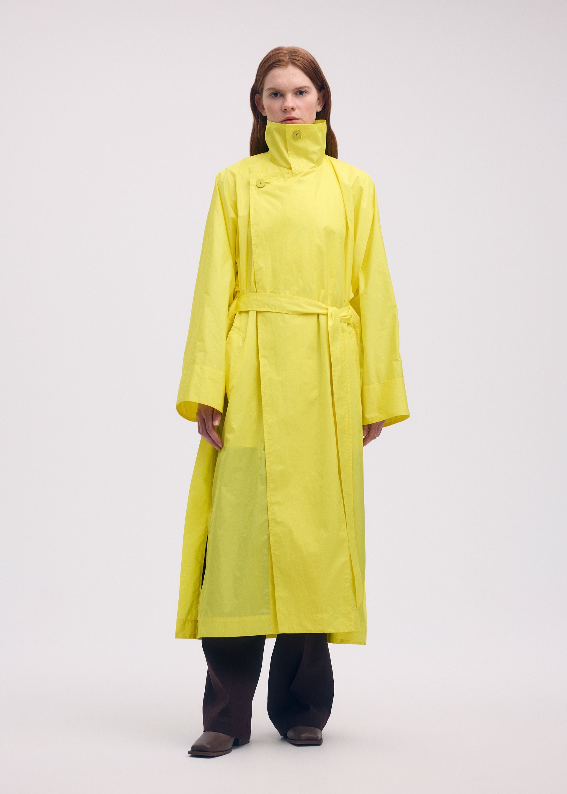 Zoomed view A person faces forward in the ISSEY MIYAKE AERIAL STEEL: a long, bright yellow lightweight taffeta coat with high collar and belt, worn over dark wide-leg pants, set against a plain light background.