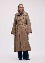 A person with shoulder-length hair wears ISSEY MIYAKE’s AERIAL STEEL, a long, loose-fitting lightweight taffeta coat with a belt, paired with black pants and shoes. The coat’s elegant luster adds sophistication against a plain light background.