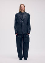 A person faces forward, wearing the ISSEY MIYAKE AERIAL STEEL: an oversized black textured blazer and wide-leg black pants with a large pocket and zipper. The background is plain and light-colored.