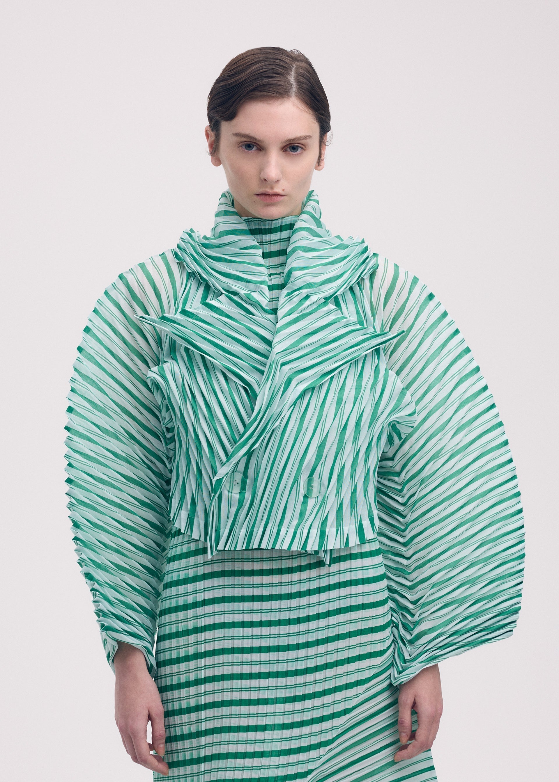 Zoomed view A person stands against a plain background, wearing the ISSEY MIYAKE GENERIC STRIPES (HAND PLEATS) set in green and white, featuring wide structured sleeves, a high collar, and a matching patterned skirt. Their hair is neatly styled back.