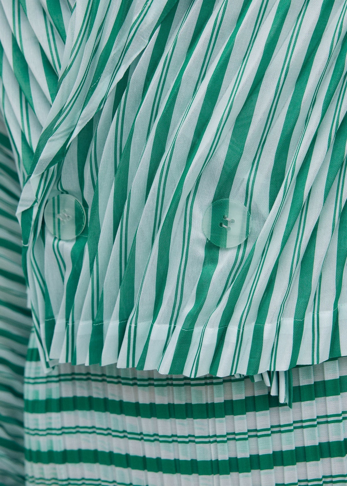 Close-up of the ISSEY MIYAKE GENERIC STRIPES (HAND PLEATS) jacket, featuring green and white diagonal stripes on top, horizontal stripes on the bottom, two translucent buttons, and sheer polyester fabric. view 5 of 5