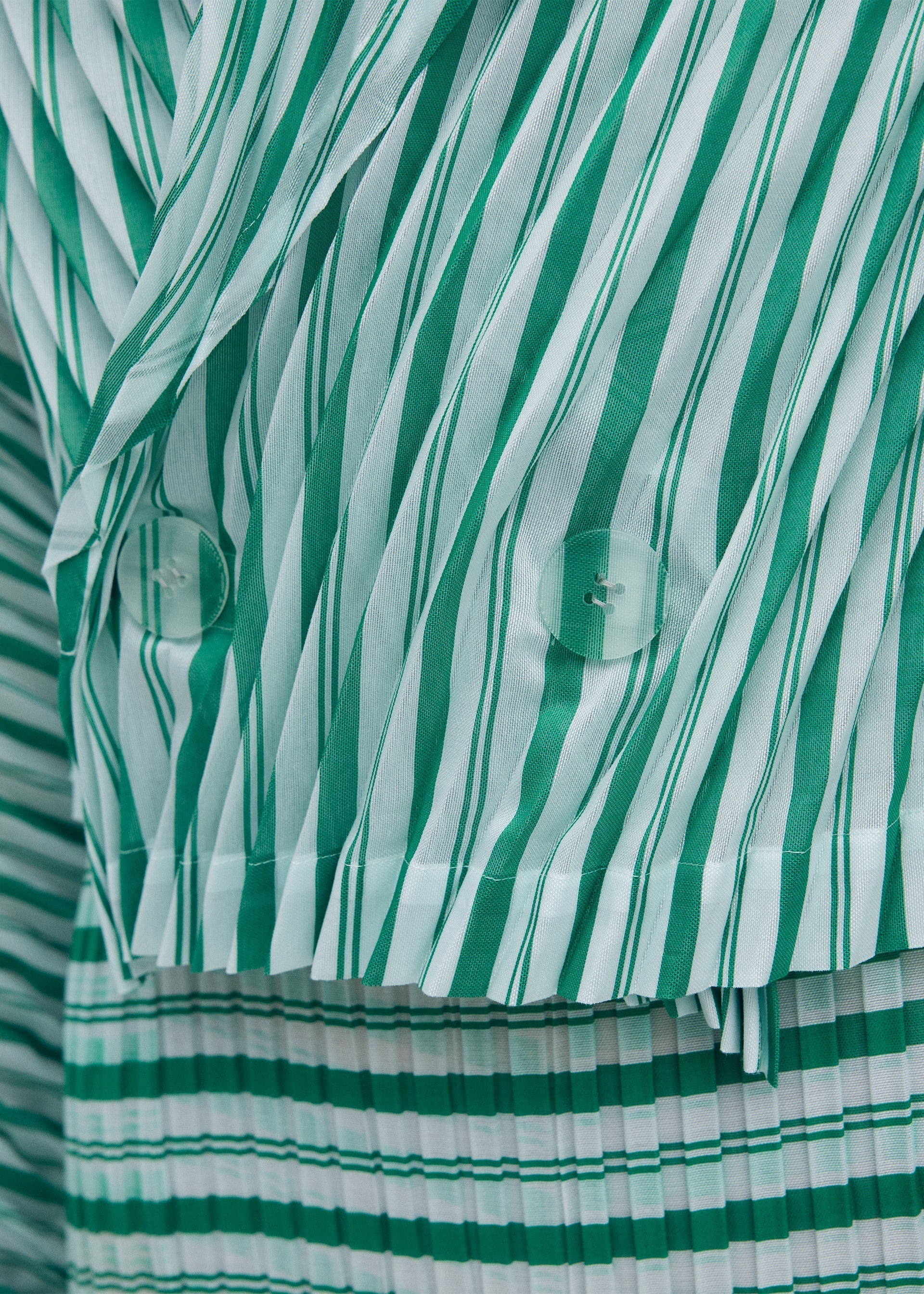 Zoomed view Close-up of the ISSEY MIYAKE GENERIC STRIPES (HAND PLEATS) jacket, featuring green and white diagonal stripes on top, horizontal stripes on the bottom, two translucent buttons, and sheer polyester fabric.