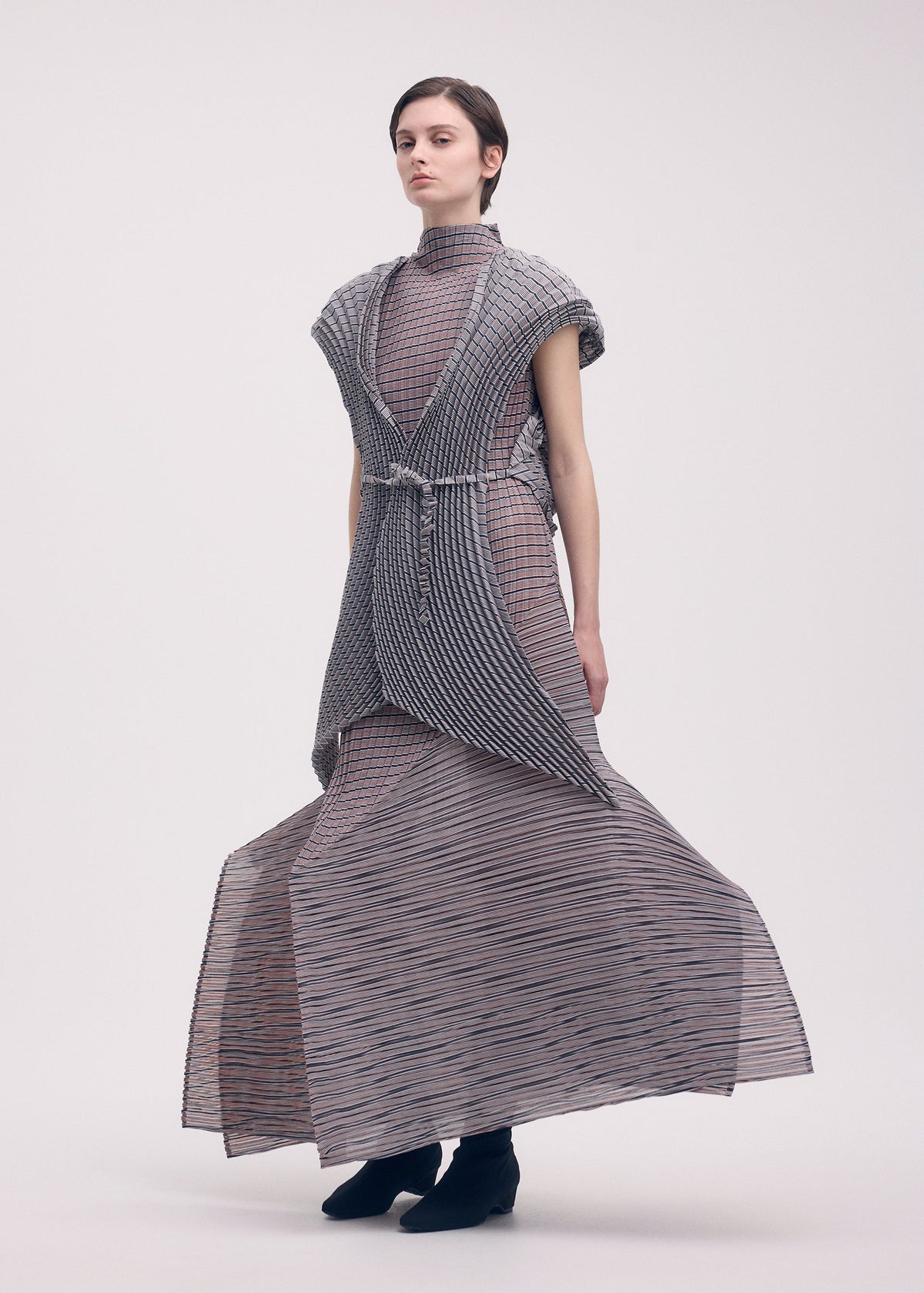 A person wears the ISSEY MIYAKE GENERIC STRIPES (HAND PLEATS) dress, featuring a high collar, floor-length handmade pleats, layered sheer striped polyester, and short sleeves, paired with black ankle boots against a plain light background. view 4 of 5