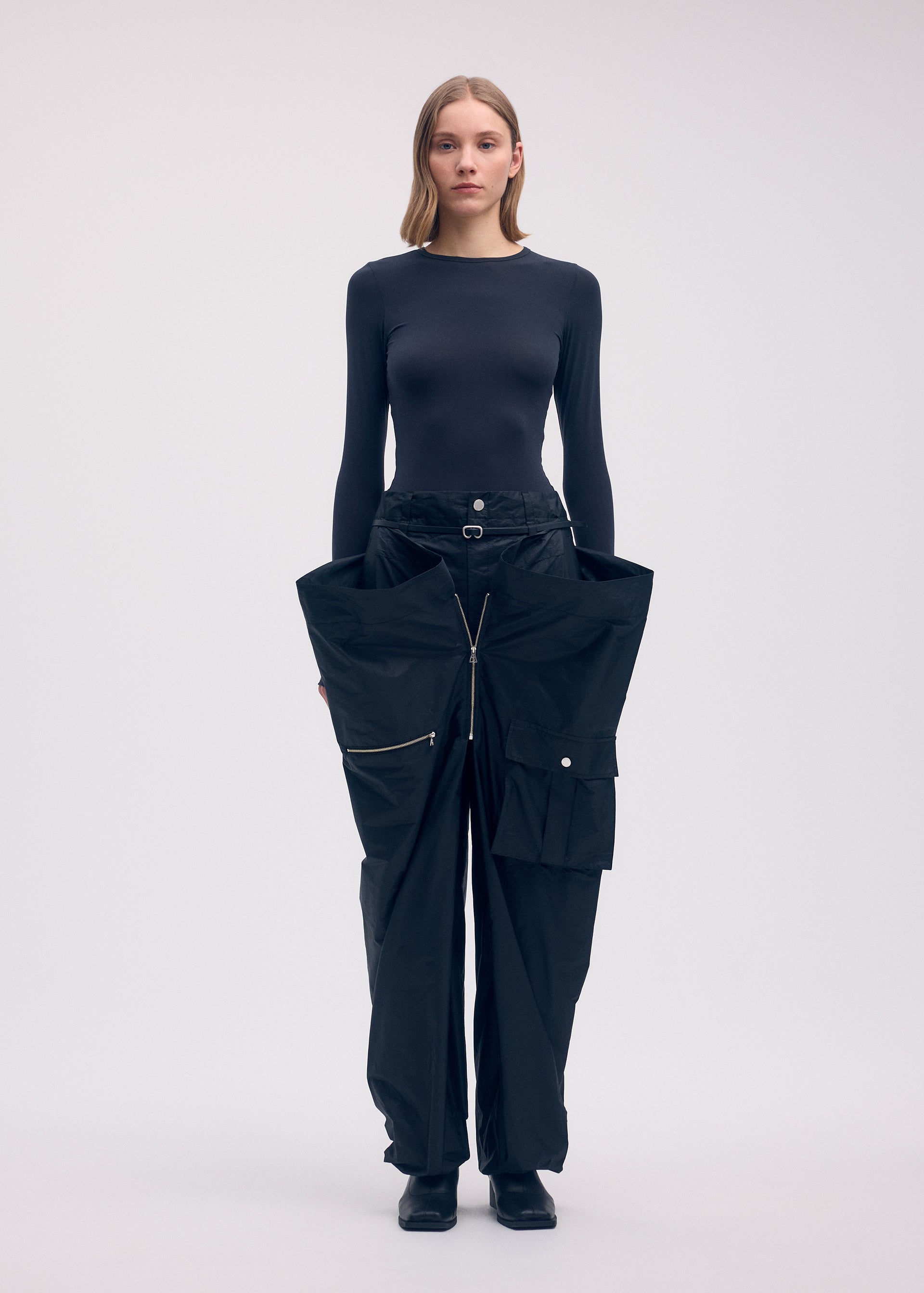 Zoomed view A woman faces forward in a fitted black top and oversized AERIAL STEEL taffeta pants by ISSEY MIYAKE, with large pockets, zippers, and a unique wide silhouette, set against a plain light background.