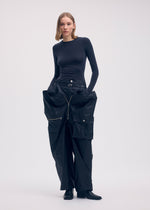A woman wears the ISSEY MIYAKE AERIAL STEEL: a fitted black long-sleeve top and wide-leg, lightweight taffeta pants with large pockets and zippers. She stands against a plain background with shoulder-length hair and black boots.