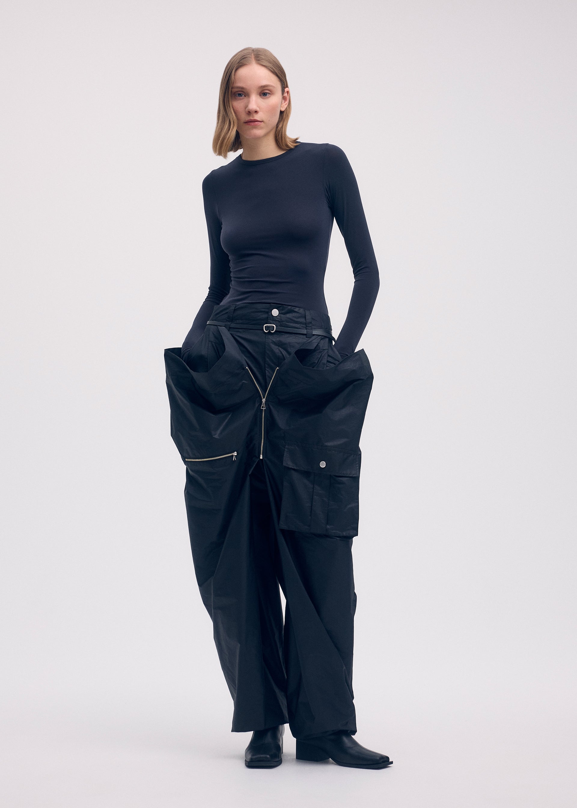 Zoomed view A woman wears the ISSEY MIYAKE AERIAL STEEL: a fitted black long-sleeve top and wide-leg, lightweight taffeta pants with large pockets and zippers. She stands against a plain background with shoulder-length hair and black boots.