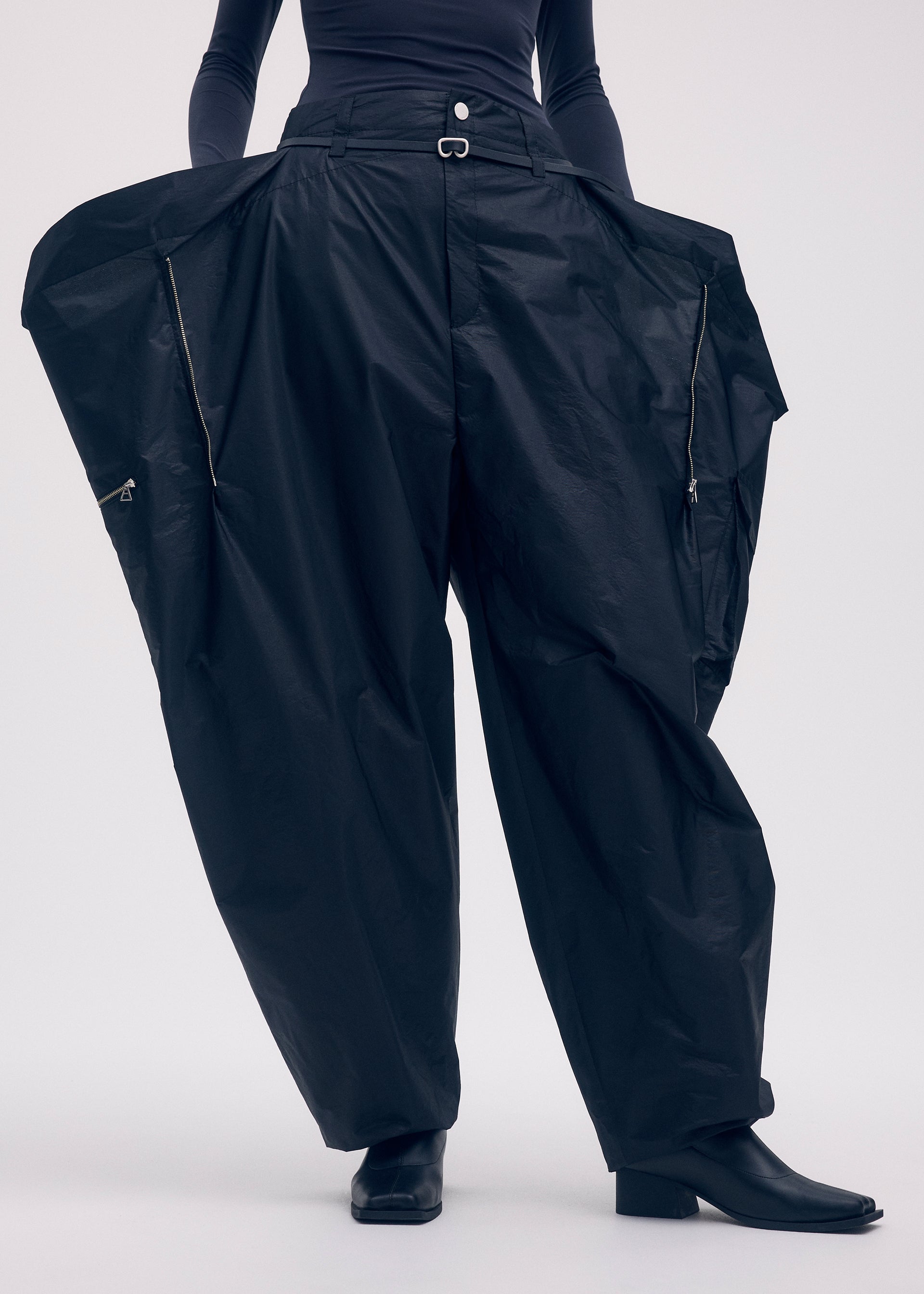 Zoomed view A person models ISSEY MIYAKE's AERIAL STEEL: voluminous black taffeta wide-leg pants with zippers, styled with black boots and a fitted top, posing against a plain background.