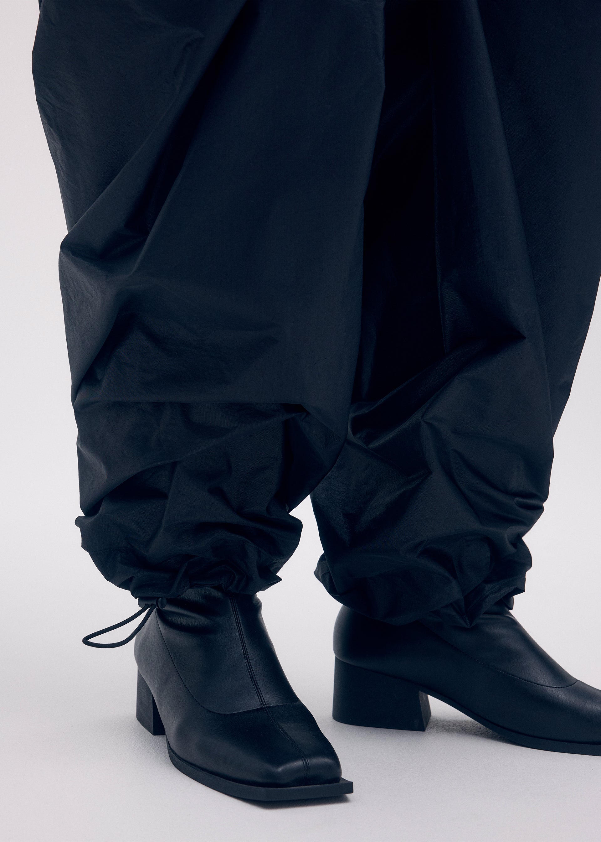 Zoomed view Close-up of a person wearing ISSEY MIYAKE AERIAL STEEL taffeta pants with an elegant luster, tucked into black square-toe block-heel ankle boots. Only the lower legs and feet are visible against a plain background.