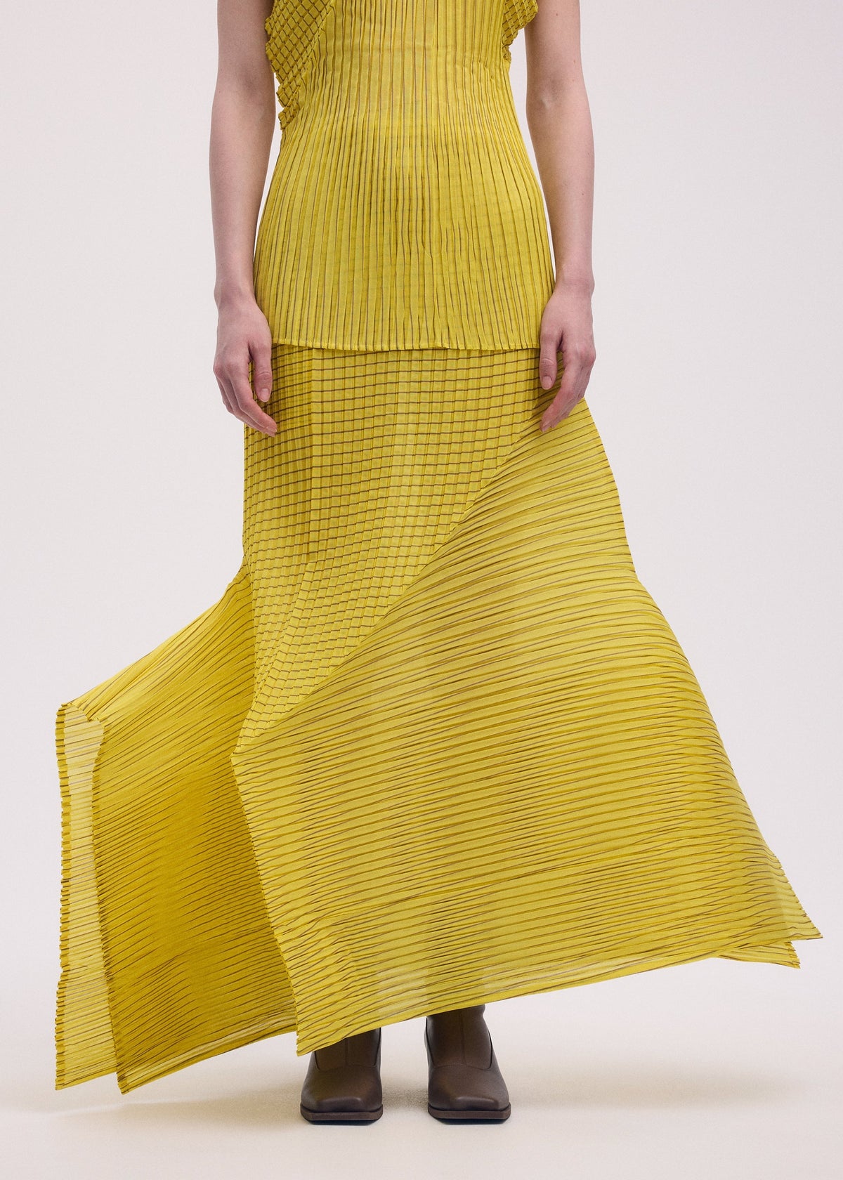 Wearing ISSEY MIYAKE’s GENERIC STRIPES (MACHINE PLEATS) yellow sleeveless top and matching pleated skirt with a moiré pattern, only the lower face and body are visible. The look is finished with brown ankle boots. view 7 of 10