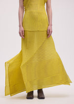 Wearing ISSEY MIYAKE’s GENERIC STRIPES (MACHINE PLEATS) yellow sleeveless top and matching pleated skirt with a moiré pattern, only the lower face and body are visible. The look is finished with brown ankle boots.
