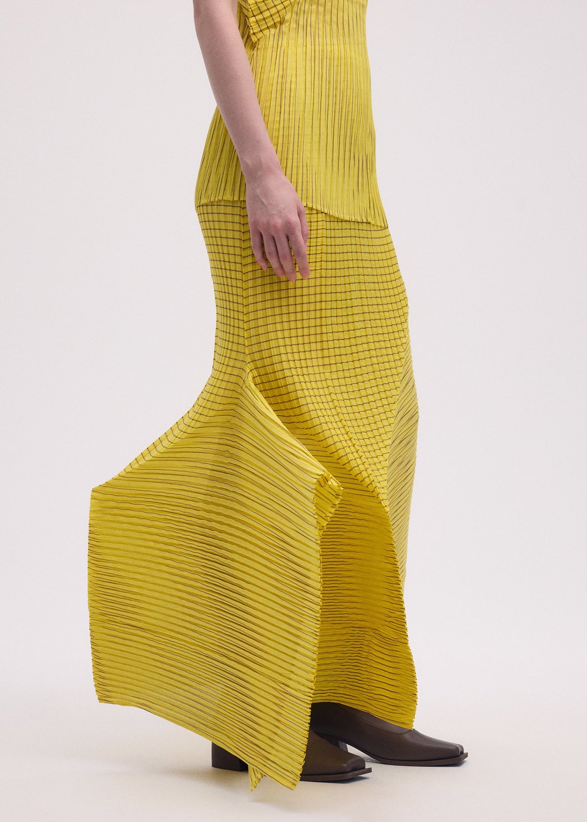 A person showcases the ISSEY MIYAKE GENERIC STRIPES (MACHINE PLEATS) yellow, striped pleated skirt with a matching top and brown heeled shoes, standing against a plain light background. Only the lower body and part of one arm are visible. view 8 of 10