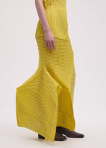 A person showcases the ISSEY MIYAKE GENERIC STRIPES (MACHINE PLEATS) yellow, striped pleated skirt with a matching top and brown heeled shoes, standing against a plain light background. Only the lower body and part of one arm are visible.