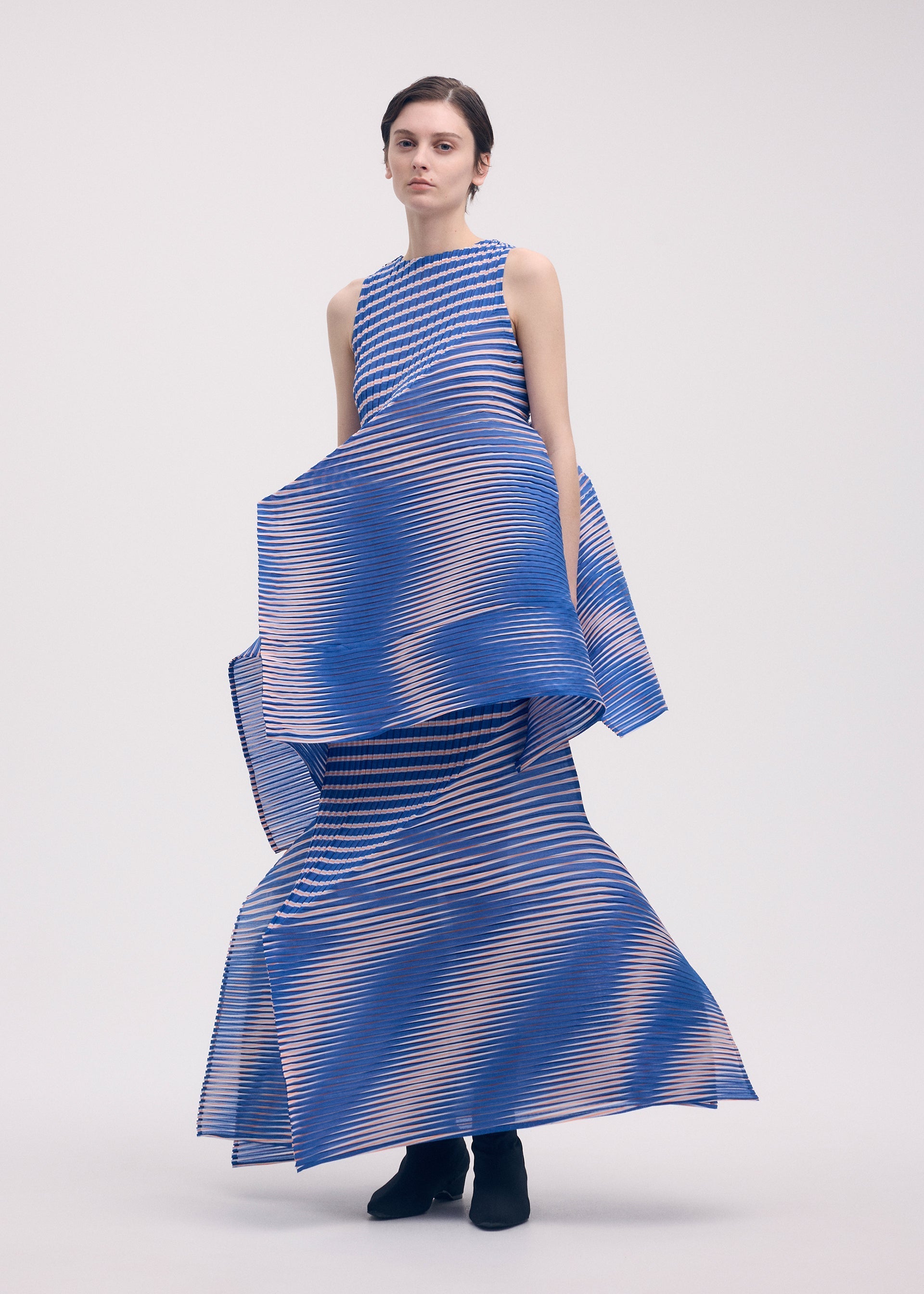 Zoomed view A woman with short dark hair wears black ankle boots and the ISSEY MIYAKE GENERIC STRIPES (MACHINE PLEATS) dress—a sleeveless, floor-length design with bold blue, white, and orange stripes and a pleated skirt—against a plain background.