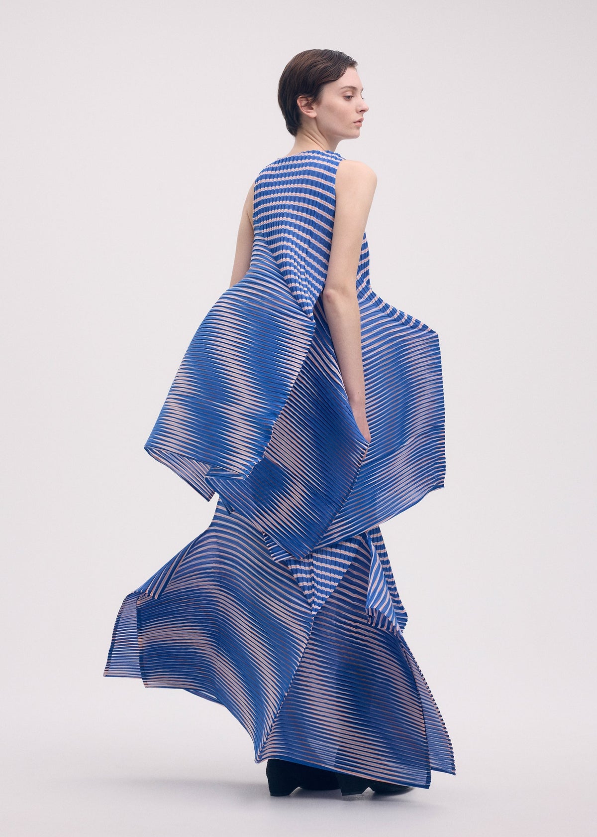 A model in the ISSEY MIYAKE GENERIC STRIPES (MACHINE PLEATS) dress, featuring geometric blue and beige stripes with sculptural pleated panels, stands sideways against a plain light background with a calm expression. view 10 of 10