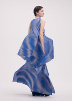 A model in the ISSEY MIYAKE GENERIC STRIPES (MACHINE PLEATS) dress, featuring geometric blue and beige stripes with sculptural pleated panels, stands sideways against a plain light background with a calm expression.