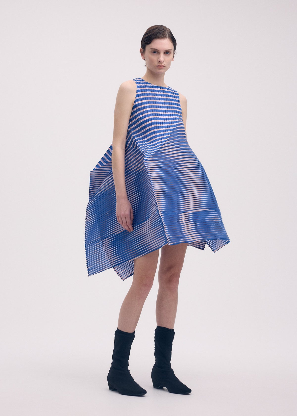 A woman models the ISSEY MIYAKE GENERIC STRIPES (MACHINE PLEATS) dress, featuring blue and white stripes with an angular, voluminous silhouette. She pairs it with black ankle boots and stands against a plain light background. view 5 of 8
