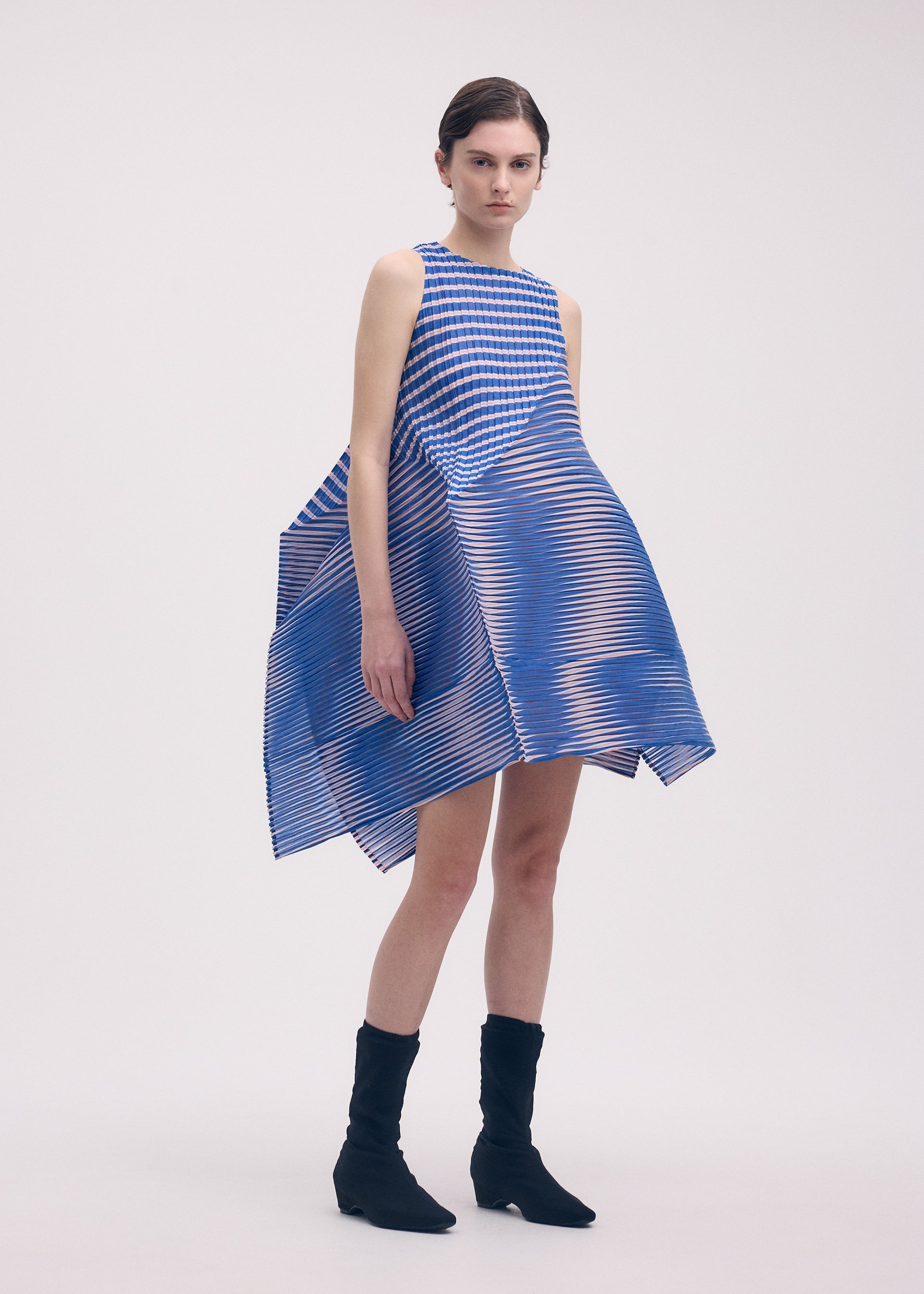Zoomed view A woman models the ISSEY MIYAKE GENERIC STRIPES (MACHINE PLEATS) dress, featuring blue and white stripes with an angular, voluminous silhouette. She pairs it with black ankle boots and stands against a plain light background.