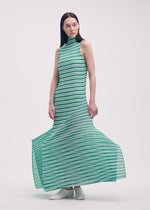 A woman with straight brown hair models the ISSEY MIYAKE GENERIC STRIPES (MACHINE PLEATS) sleeveless maxi dress with green and white stripes and elegant pleats, paired with white shoes, against a plain light background.