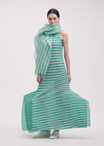 A woman stands against a white background wearing the ISSEY MIYAKE GENERIC STRIPES (MACHINE PLEATS) sleeveless dress with green and white stripes, a matching oversized scarf, and stylish white ankle boots.