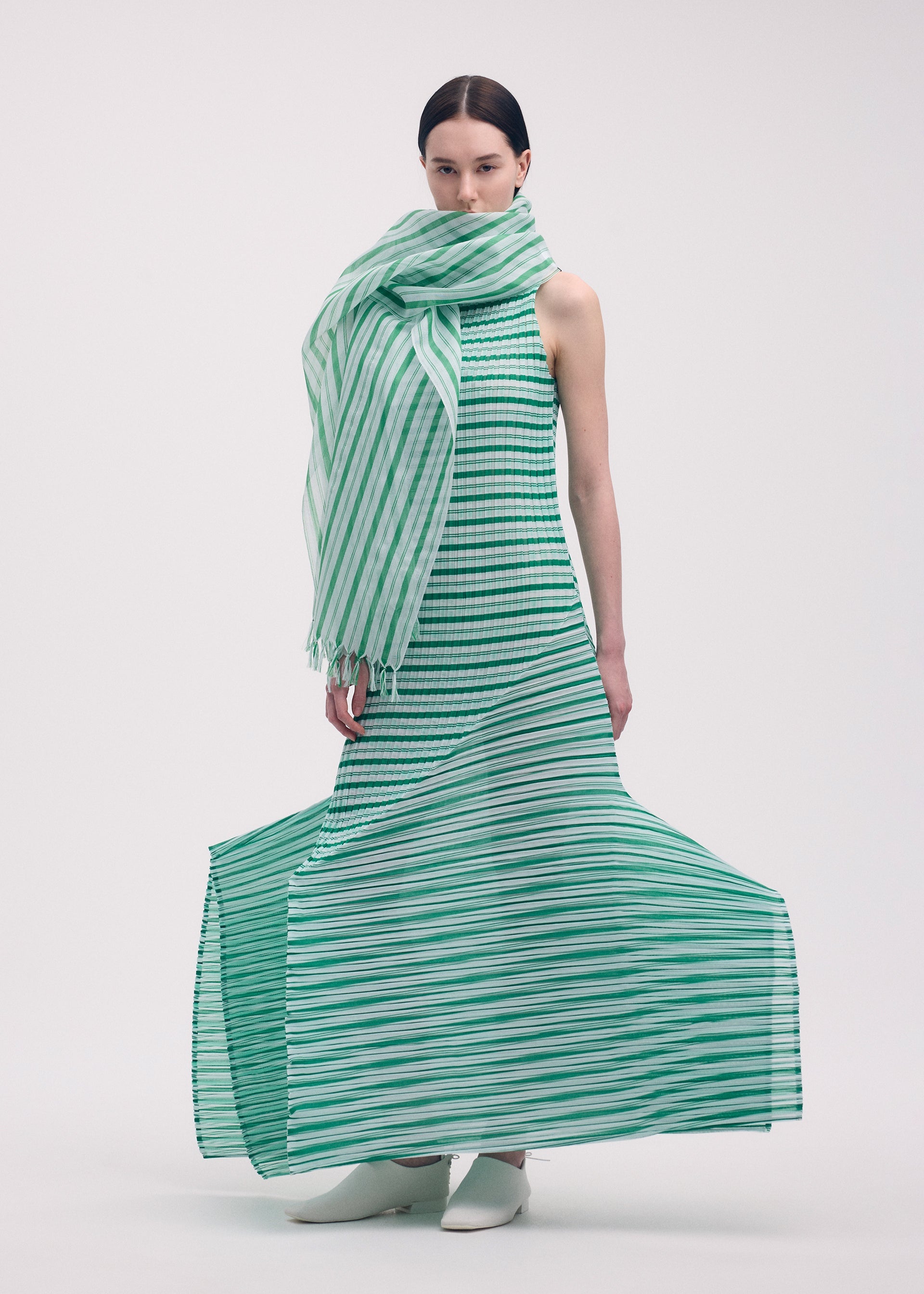 Zoomed view A woman stands against a white background wearing the ISSEY MIYAKE GENERIC STRIPES (MACHINE PLEATS) sleeveless dress with green and white stripes, a matching oversized scarf, and stylish white ankle boots.
