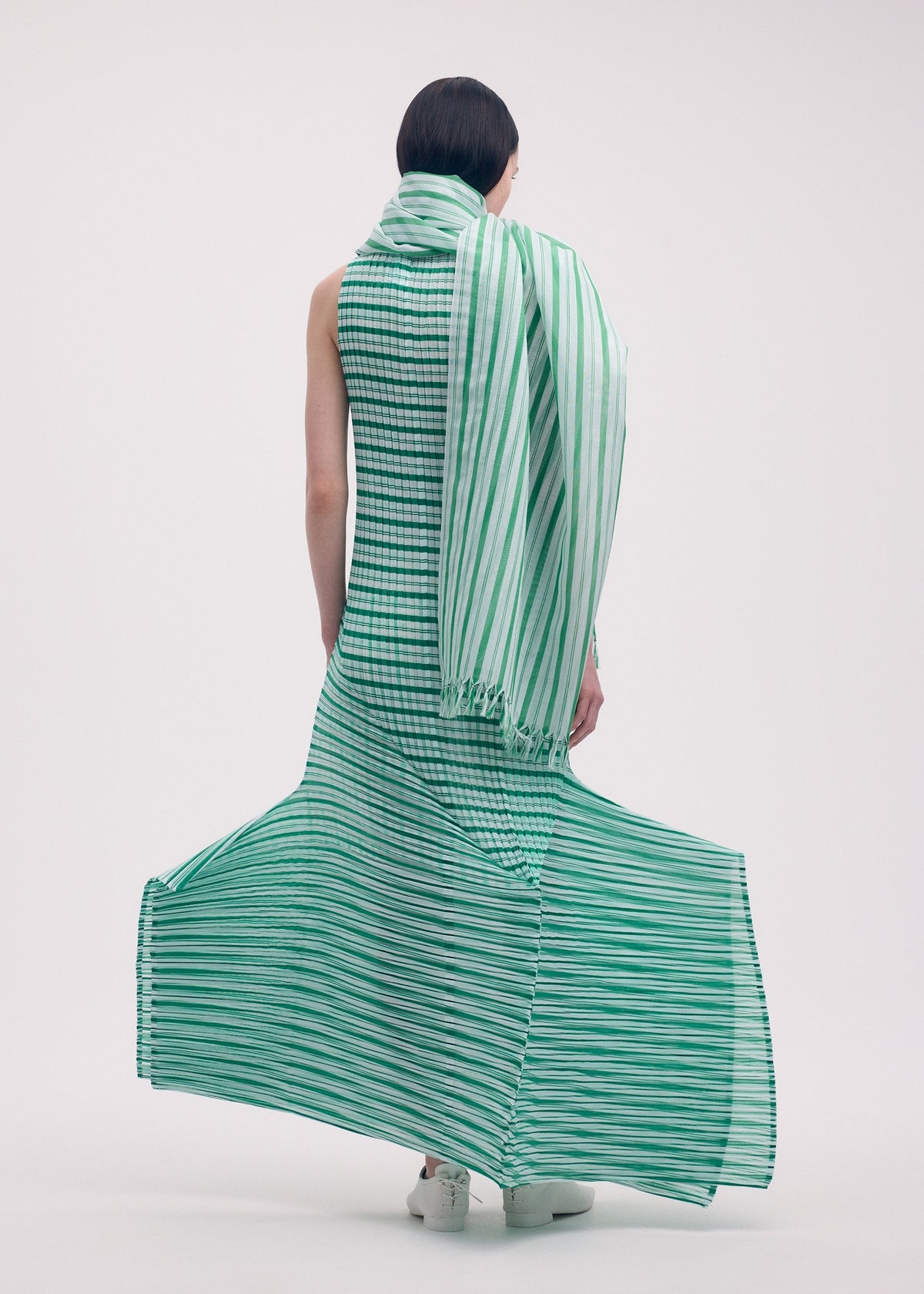 A person with short dark hair models the ISSEY MIYAKE GENERIC STRIPES (MACHINE PLEATS) dress—long, sleeveless, green-and-white striped with a matching scarf—standing against a plain white background as the hem flares in soft pleats. view 6 of 6
