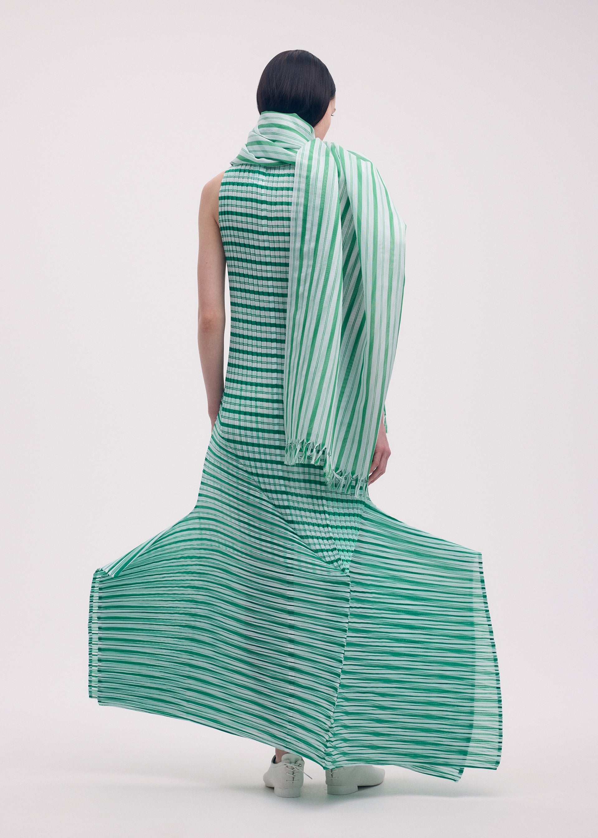 Zoomed view A person with short dark hair models the ISSEY MIYAKE GENERIC STRIPES (MACHINE PLEATS) dress—long, sleeveless, green-and-white striped with a matching scarf—standing against a plain white background as the hem flares in soft pleats.