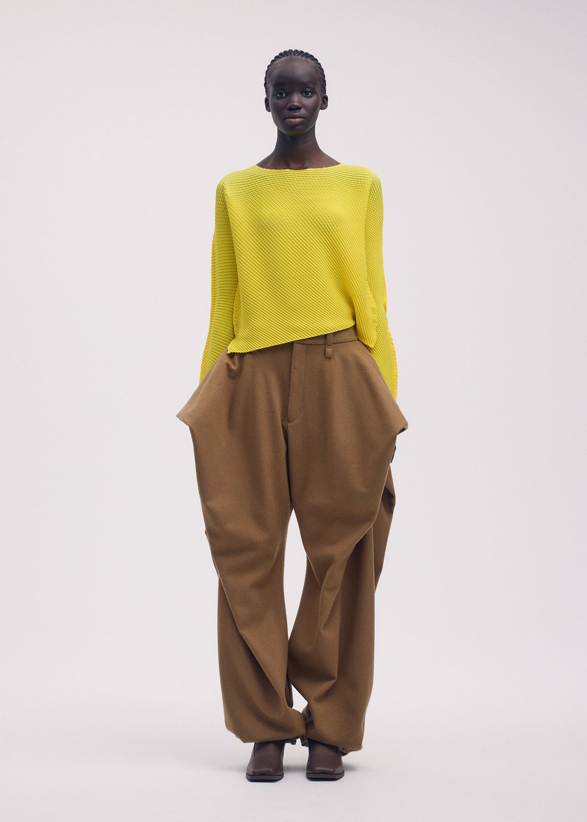 A person wears the ISSEY MIYAKE SHIFTED MISTY PLEATS: a striking yellow long-sleeved top with unique design, paired with loose brown pants featuring wide, structured hips and dark shoes, standing against a plain background. view 6 of 7